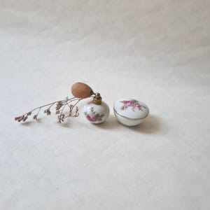 Op de afbeelding: Een set van twee porseleinen parfumflesjes met bloemenmotieven. De flesjes zijn wit met roze en groene bloemenpatronen. Het kleinere flesje heeft een gouden deksel en een bruine sproeikop. Het grotere flesje heeft een rond deksel met een gouden rand.