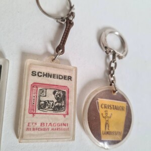 May include: Two vintage keychains. One is a clear plastic keychain with a red and white image of a television with a teddy bear inside. The text on the keychain reads "Schneider Ets Biaggini 81, B.P.Chave Marseille". The other keychain is a clear plastic keychain with a brown and yellow image of a man holding a bucket with the text "Cristalor  L'Anoussi&egrave;re".
