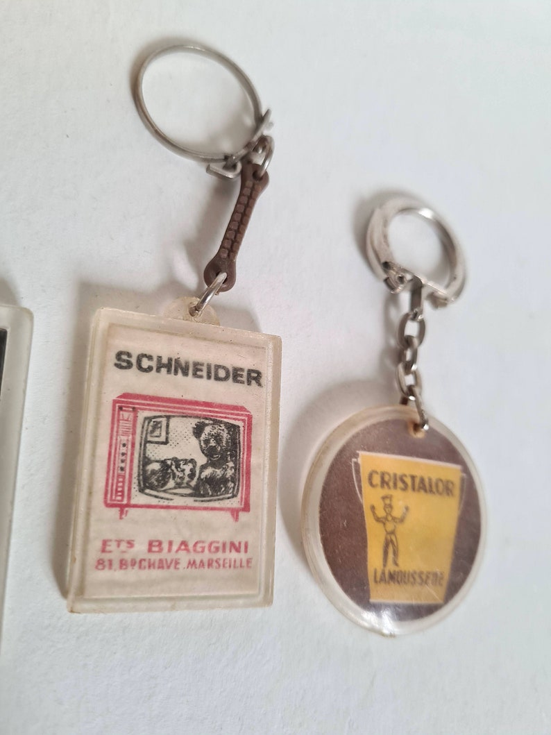 May include: Two vintage keychains. One is a clear plastic keychain with a red and white image of a television with a teddy bear inside. The text on the keychain reads "Schneider Ets Biaggini 81, B.P.Chave Marseille". The other keychain is a clear plastic keychain with a brown and yellow image of a man holding a bucket with the text "Cristalor  L'Anoussi&egrave;re".