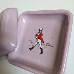 K&ouml;nnte beinhalten: Ein quadratischer Aschenbecher in helllila mit einem Johnnie Walker Logo. Das Logo zeigt einen Mann in roter Jacke und Zylinder, der mit einem Stock geht. Der Text "Born 1820" und "Still Going Strong" ist unter dem Mann gedruckt.