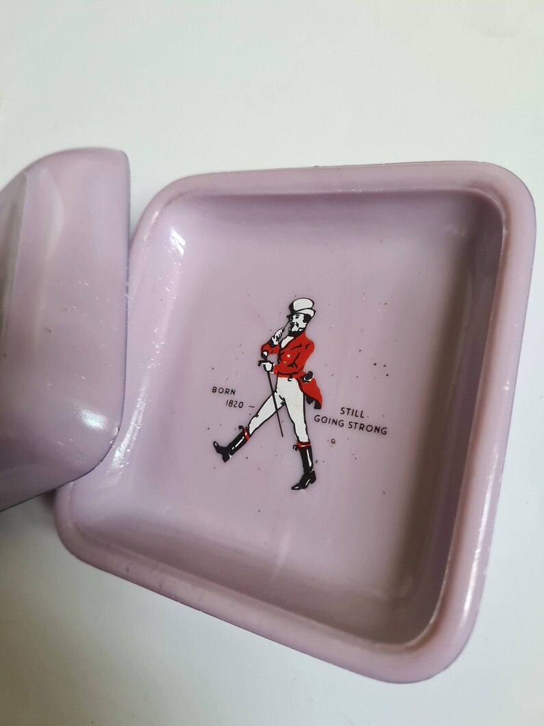 K&ouml;nnte beinhalten: Ein quadratischer Aschenbecher in helllila mit einem Johnnie Walker Logo. Das Logo zeigt einen Mann in roter Jacke und Zylinder, der mit einem Stock geht. Der Text "Born 1820" und "Still Going Strong" ist unter dem Mann gedruckt.