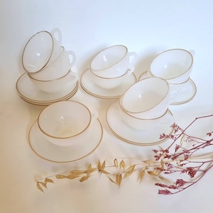 May include: Set of six white teacups and saucers with gold trim. The cups and saucers are stacked and arranged on a white surface.