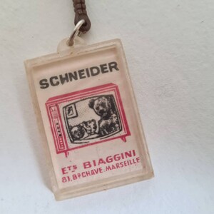 May include: A white plastic keychain with a red and black image of a television set with two dogs inside. The text "SCHNEIDER" is printed in black above the television and "ETS BIAGGINI 81, BPCHAVE.MARSEILLE" is printed in red below the television.