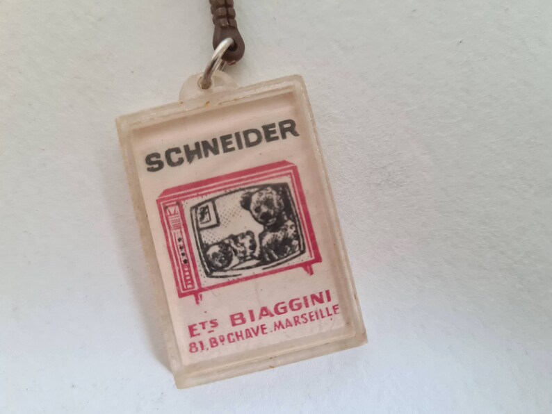 May include: A white plastic keychain with a red and black image of a television set with two dogs inside. The text "SCHNEIDER" is printed in black above the television and "ETS BIAGGINI 81, BPCHAVE.MARSEILLE" is printed in red below the television.
