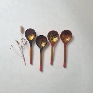 Peut inclure: Quatre cuillères en bois avec des motifs floraux ornés. Les cuillères ont des cuillères noires avec des accents rouges et dorés. Les manches sont en bois naturel avec des pointes rouges. Une brindille de fleurs séchées est à gauche des cuillères.