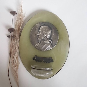 May include: Oval-shaped green stone plaque with a silver-toned relief of Jesus Christ's face. The plaque has a small silver-toned rectangular plate with a floral design and a glass vial.