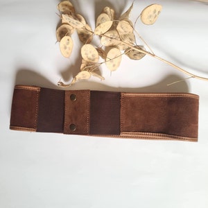 Può includere: Una cintura in pelle scamosciata marrone con un inserto in tessuto marrone e due bottoni metallici.