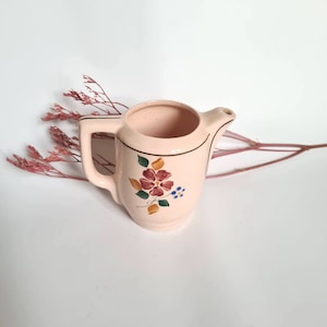 May include: A small pink ceramic creamer with a floral design. The creamer has a spout and a handle. The flowers are red, yellow, blue, and green.