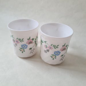 May include: Two white, cylindrical cups with a floral design. The cups feature pink, blue, and gray flowers with green leaves. The cups are empty and sit on a light-colored surface.