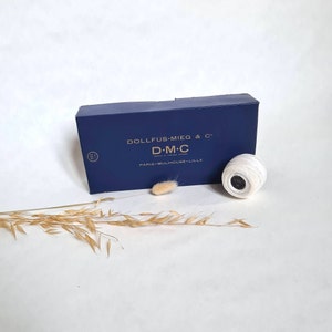 May include: A blue box with gold lettering that reads "DOLLFUS-MIEG & C" and "D-M-C" with the text "PARIS-MULHOUSE-LILLE" below. A white ball of thread with a white label that reads "DMC" is sitting on a white surface next to the box.
