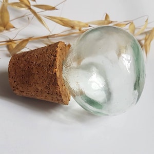 May include: A clear glass bottle stopper with a cork base. The bottle stopper is round and has a slight green tint.