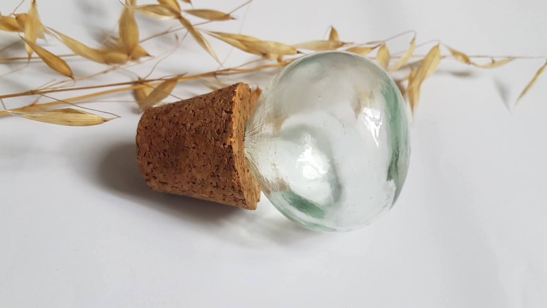 May include: A clear glass bottle stopper with a cork base. The bottle stopper is round and has a slight green tint.