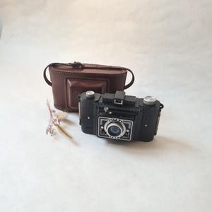 May include: A vintage black Ultra camera with a silver lens and dial, accompanied by a brown leather case. The camera has a rectangular shape with a silver frame around the lens. A small sprig of dried flowers sits nearby.