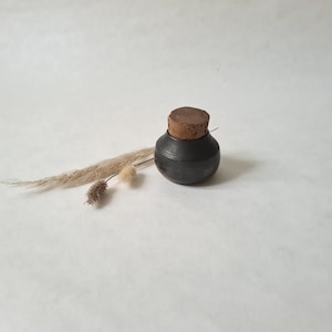 Peut inclure: Un petit pot en céramique noire avec un couvercle en liège. Le pot est posé sur une surface blanche.