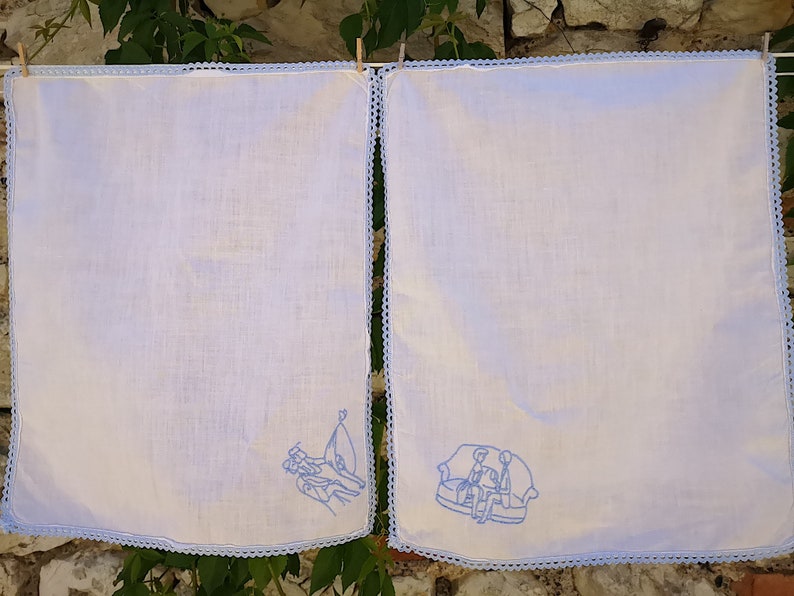 Puede incluir: Dos toallas de cocina blancas con dise&ntilde;os bordados en azul. Una toalla presenta un grupo de personas de pie, la otra presenta un grupo de personas sentadas en un sof&aacute;.