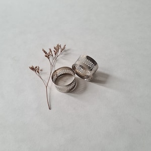 May include: Two silver napkin rings with a geometric design. The rings are on a white surface.