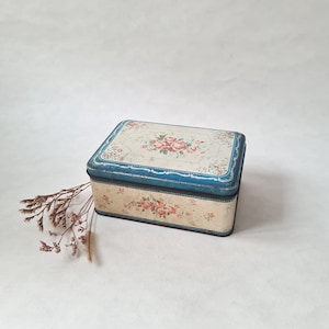 May include: A vintage rectangular tin box with a floral pattern. The box has a cream base with pink roses and green leaves. The lid and trim are a faded blue. A sprig of dried flowers is placed beside the box.