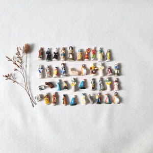 May include: A collection of small, hand-painted ceramic figurines arranged in four rows, featuring various human figures in colorful attire, alongside a single dried plant stem with thin, branching twigs positioned to the left side.