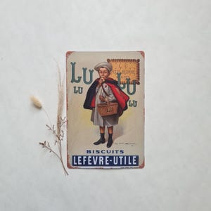 Peut inclure: Une plaque en métal vintage faisant la publicité des biscuits Lu. La plaque représente un jeune garçon en manteau rouge tenant un panier de biscuits Lu. Le texte sur la plaque est "Lu Biscuits Lefevre-Utile".