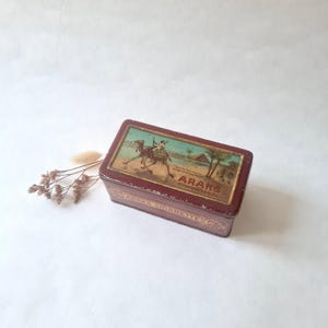 Puede incluir: Una caja de lata vintage rectangular de color granate con una tapa decorativa que presenta una escena del desierto con un camello y un jinete. La caja tiene detalles dorados y el texto "ARAKS CIGARETTES".
