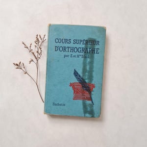 Peut inclure: Un livre vintage intitulé "COURS SUPÉRIEUR D'ORTHOGRAPHE" en français, avec une couverture bleu clair. La couverture présente une illustration de machine à écrire et de plume rouge. Le livre est légèrement usé. Une brindille de fleurs séchées est posée à côté.