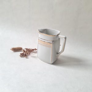 Puede incluir: Jarra de cerámica blanca con cuerpo cuadrado y asa. La jarra está decorada con ribetes dorados alrededor de la parte superior, el asa y una banda rectangular de filigrana dorada. Una ramita de flores secas descansa junto a la jarra.