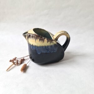 Op de afbeelding: Een keramische creamer met een gebogen handvat en tuit. De creamer heeft een kleurovergang, van beige aan de bovenkant naar bruin, blauw en uiteindelijk zwart aan de basis. Een takje gedroogde bloemen rust bij de creamer.