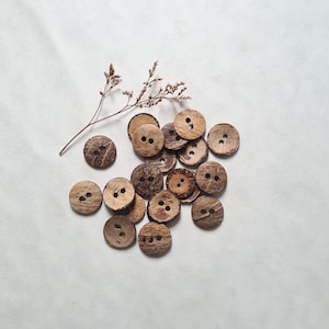 Peut inclure: Un ensemble de boutons en coquille de noix de coco brune avec deux trous chacun. Les boutons sont disposés en pile sur une surface blanche.
