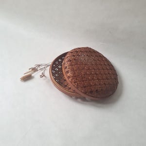 Peut inclure: Un panier rond en osier avec un couvercle, de couleur marron. Le couvercle est partiellement ouvert, révélant l'intérieur. Le panier a une surface texturée. Une brindille de fleurs séchées est à côté du panier.