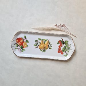 Peut inclure: Un plateau de service rectangulaire en céramique blanche avec une bordure dorée. Il est orné d'illustrations colorées de pommes, de poires et de cerises avec des accents floraux. Un objet décoratif pour servir.