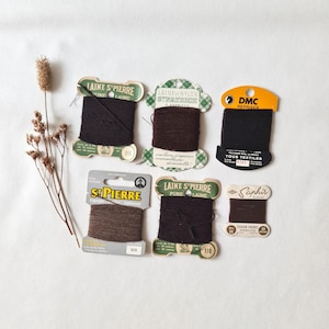 Può includere: Una collezione di sei cartelle di filo da rammendo vintage in varie tonalità di marrone e nero, con marchi come Laine St. Pierre, St. Patrick, DMC e Saphir. Le cartelle sono disposte con uno stelo botanico essiccato sul lato sinistro.
