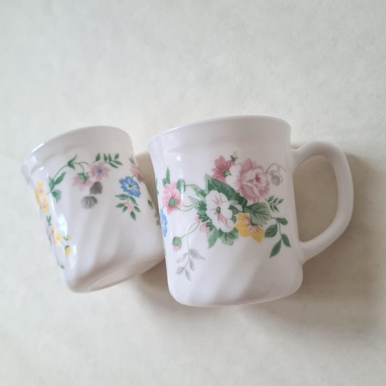 May include: Two white ceramic mugs with floral designs. The mugs feature a handle and a curved shape. The floral pattern includes pink, blue, yellow, and green flowers and leaves. The mugs are set against a light background.