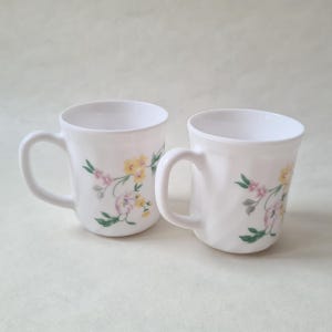 May include: Two white ceramic mugs with floral designs. Each mug features a curved handle and is decorated with yellow, pink, and green floral patterns. The mugs are set against a light background.