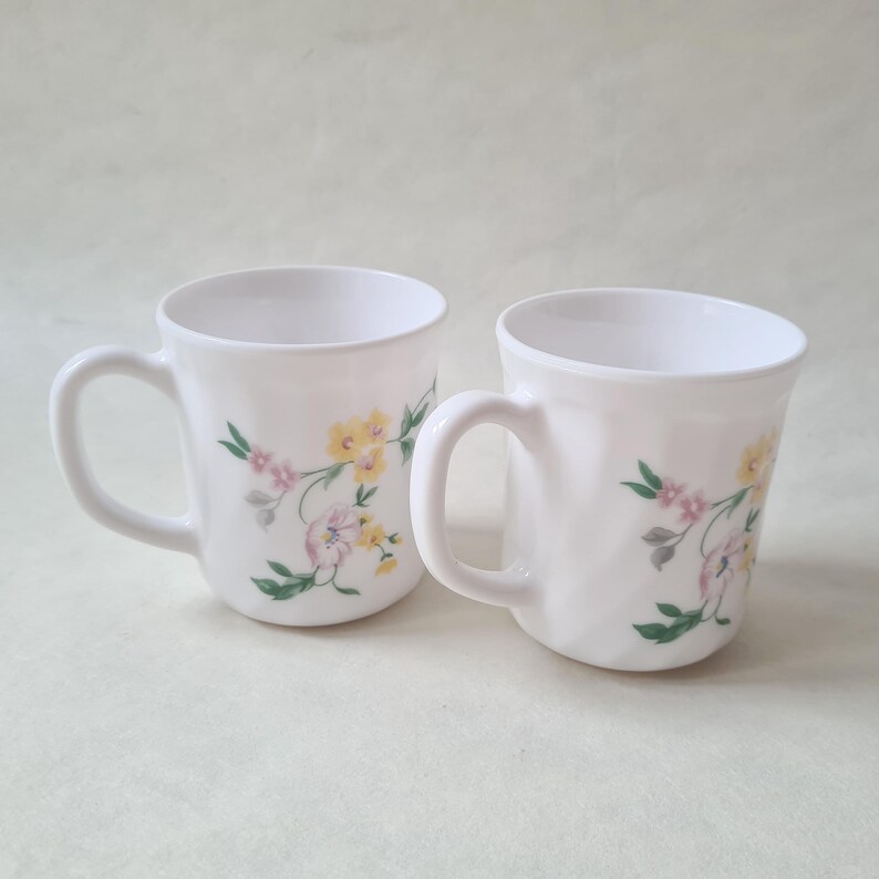 May include: Two white ceramic mugs with floral designs. Each mug features a curved handle and is decorated with yellow, pink, and green floral patterns. The mugs are set against a light background.