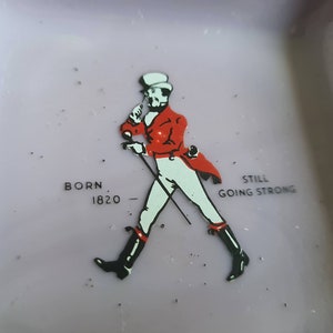 K&ouml;nnte beinhalten: Ein lilafarbener Aschenbecher aus Keramik mit einer Schwarzwei&szlig;illustration eines Mannes in roter Jacke und Zylinder. Der Text "BORN 1820" und "STILL GOING STRONG" ist auf dem Aschenbecher aufgedruckt.