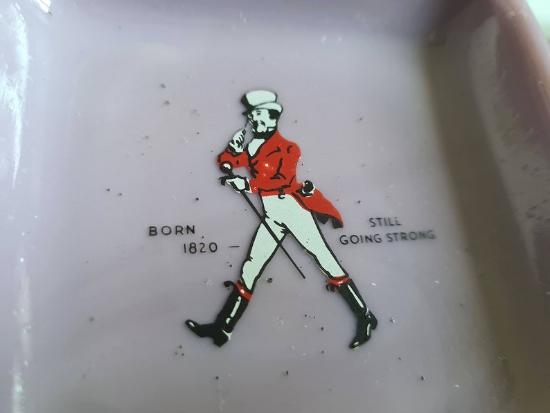 K&ouml;nnte beinhalten: Ein lilafarbener Aschenbecher aus Keramik mit einer Schwarzwei&szlig;illustration eines Mannes in roter Jacke und Zylinder. Der Text "BORN 1820" und "STILL GOING STRONG" ist auf dem Aschenbecher aufgedruckt.
