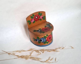 May include: A wooden salt cellar with a hinged lid, painted with colorful flowers and leaves. The cellar has a small hole on the back for hanging.