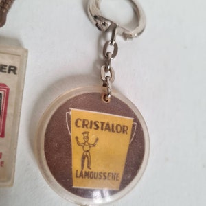 May include: A round, clear plastic keychain with a brown and yellow image of a bucket and a stick figure. The bucket says "Cristalor Lamoussette" in white text.