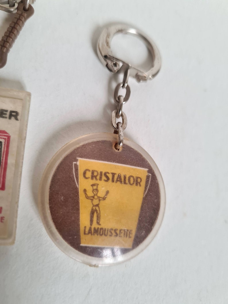 May include: A round, clear plastic keychain with a brown and yellow image of a bucket and a stick figure. The bucket says "Cristalor Lamoussette" in white text.