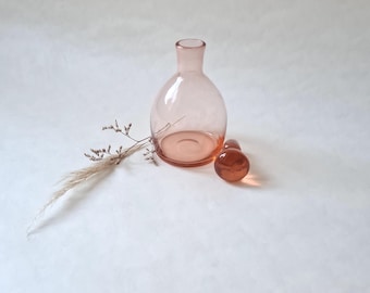 May include: A light pink glass bottle with a round body and a narrow neck. The bottle is sitting on a white surface with two small, round, light pink glass stoppers.