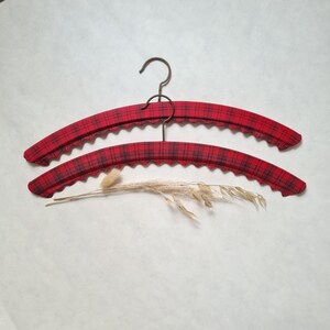 May include: Two red plaid padded clothes hangers with a scalloped edge and a bronze hook. The hangers are arranged in a layered composition, with a sprig of dried grass in the foreground.