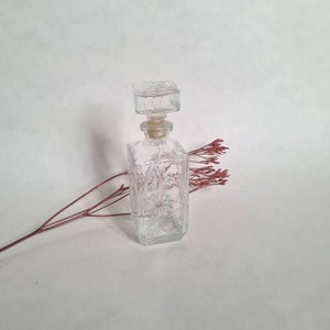May include: A clear glass bottle with a square stopper and a floral design. The bottle is empty and has a textured surface.