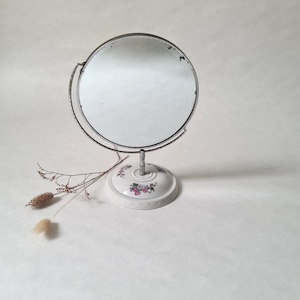 May include: A vintage vanity mirror with a round, silver-framed mirror and a white porcelain base with a floral design.
