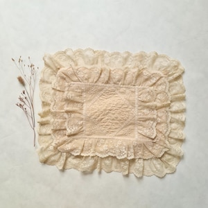 Puede incluir: Un tapete de encaje rectangular de color crema con un patrón central de diamante acolchado rodeado por múltiples capas de encaje con volantes y motivos florales, acompañado de un tallo botánico seco con pequeños capullos marrones.