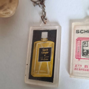 May include: A clear plastic keychain with a small bottle of cologne inside. The bottle is yellow and has the text "Eau de Cologne" and "Luxe Ambiance Werbauge" on it.