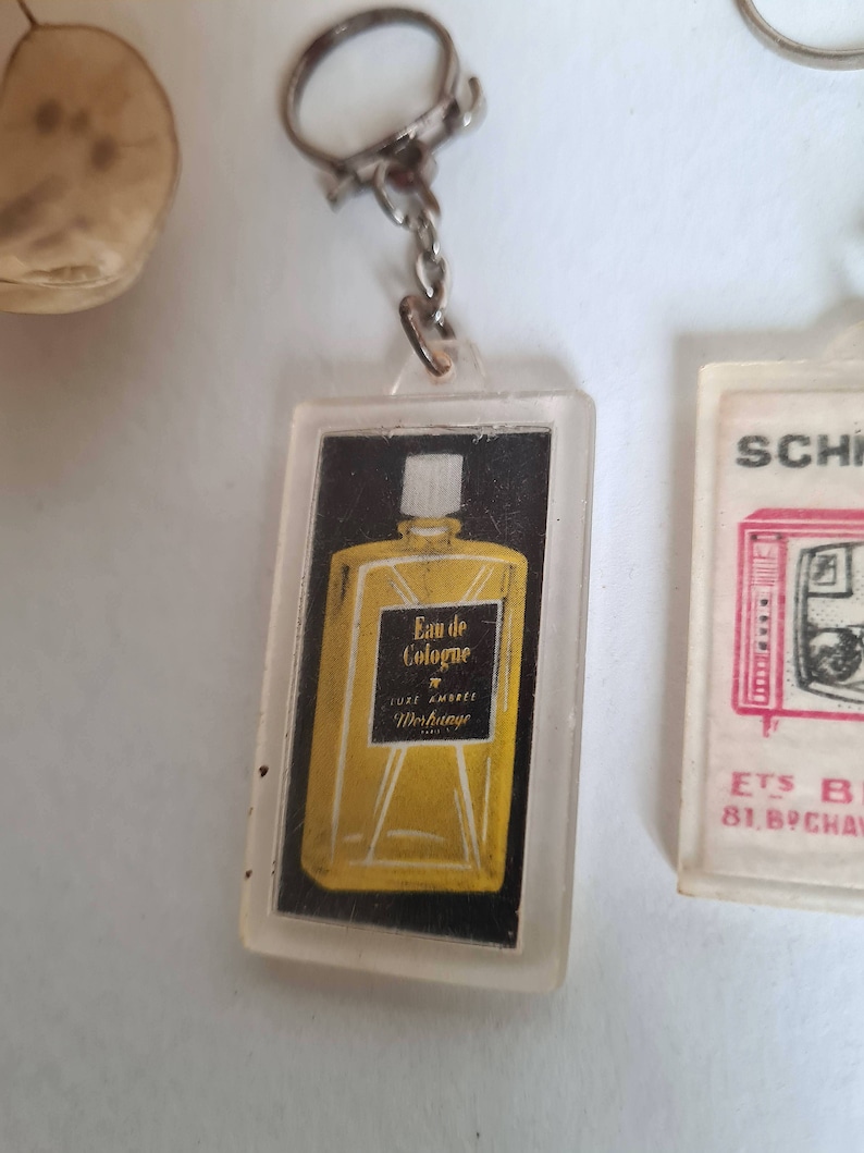 May include: A clear plastic keychain with a small bottle of cologne inside. The bottle is yellow and has the text "Eau de Cologne" and "Luxe Ambiance Werbauge" on it.