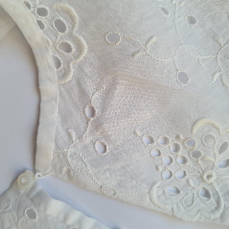 May include: White cotton fabric with a delicate eyelet embroidery pattern. The fabric has a soft, textured feel and is perfect for creating garments or home decor.