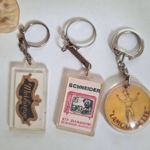 May include: Three vintage keychains with different designs. The first keychain features a brown and black logo with the text "Moskava". The second keychain has a red and white image of a television with the text "Schneider" and "Ets. Biaggini 61, Bd.G.Chave Marseille". The third keychain has a yellow background with a cartoon image of a person in a red and white outfit with the text "L'Amour Petite".