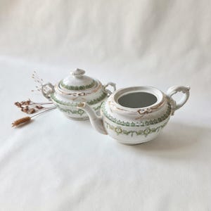 Könnte beinhalten: Eine weiße Porzellan-Zuckerdose mit Deckel und ein passendes Milchkännchen, beide mit einem grünen und goldenen Dekorationsmuster mit Schleifenmotiven. Das Set umfasst zwei Henkel an der Zuckerdose sowie einen Henkel und Ausguss am Kännchen.