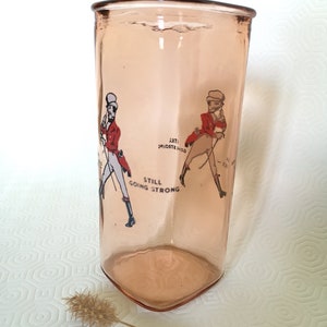 Peut inclure: Un verre en verre rose avec un design vintage repr&eacute;sentant deux hommes en veste rouge et pantalon blanc. Le texte "Still Going Strong" est imprim&eacute; sur le verre.